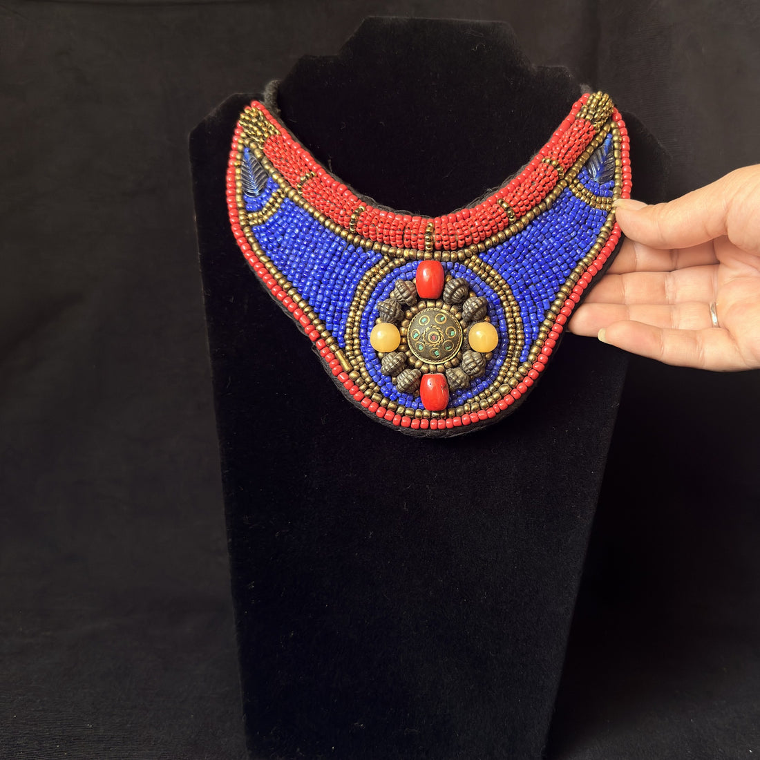 A hand wearing a silver bracelet holds a colorful beaded collar necklace against a black velvet display bust. The necklace features a vibrant blue center, red beaded borders, and gold and red beadwork details.