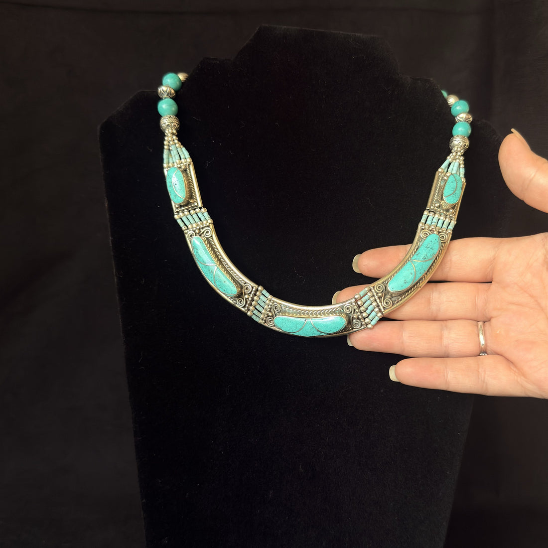 A silver-toned statement necklace with turquoise-colored stone inlays, displayed on a black velvet jewelry bust with a person's hand holding the right side of the necklace for scale.