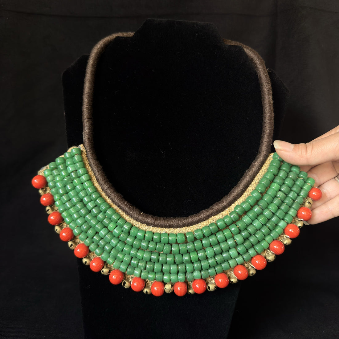 A wide, multi-strand statement necklace displayed on a black velvet mannequin bust, with a hand holding the right side to show its flexibility. The necklace features multiple rows of textured green beads, a brown braided cord trim, and a border of small red-orange beads and gold-toned spacer beads.