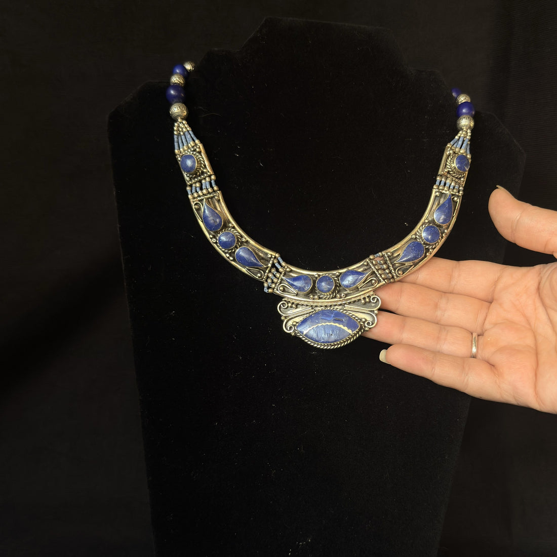 A silver-toned statement necklace with blue stone inlays and a central teardrop pendant, displayed on a black velvet stand with a person's hand reaching toward it against a black background.