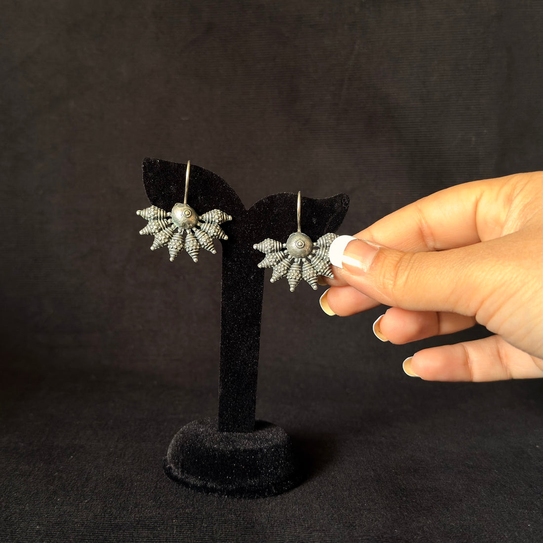 A pair of silver-toned dangle earrings with textured sunburst designs and central pearl-like beads, displayed on a black stand while a hand holds one earring against a dark background.